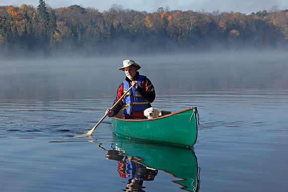 Man Paddling a Canoe with a Small White Dog in the Bow